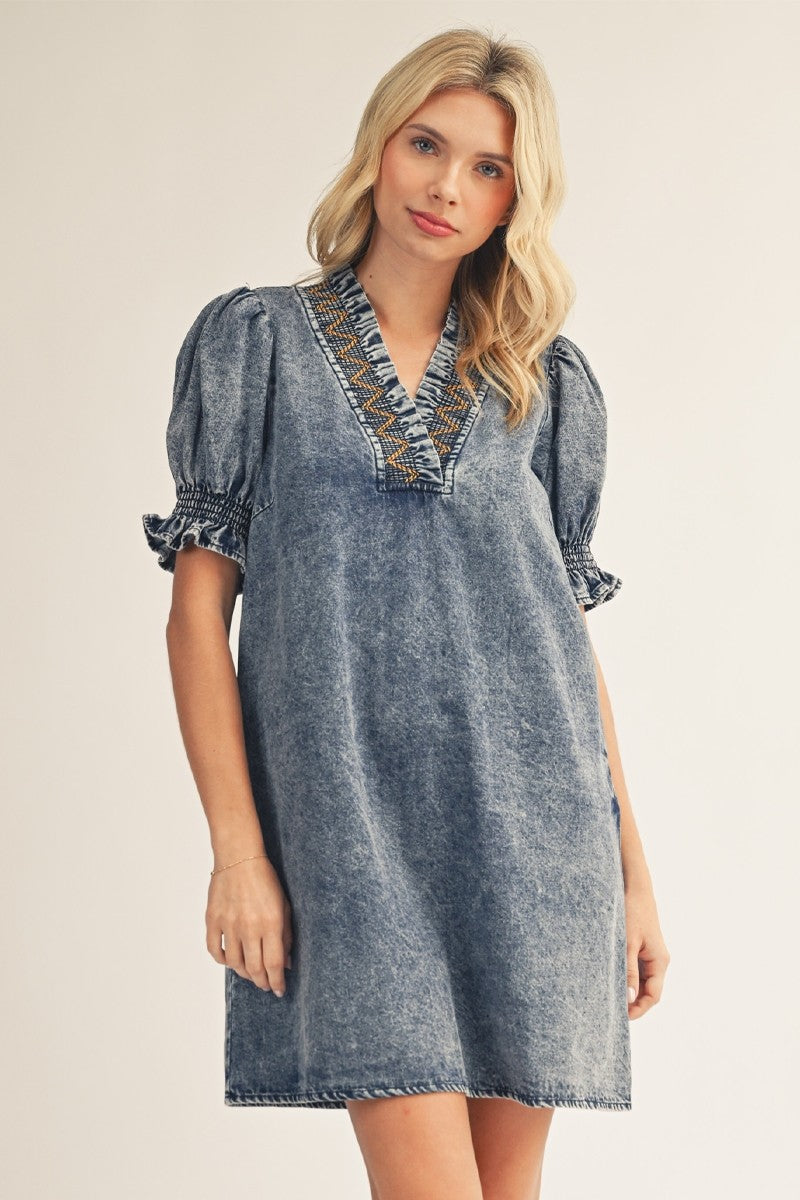 Woman wearing a denim dress with embroidered neckline on a plain background