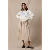 Woman wearing a white sweater with a blue bow design and beige skirt against a beige background
