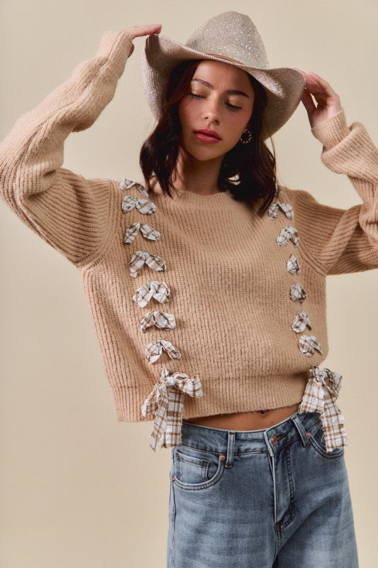 Woman wearing a beige sweater with bow details, blue jeans, and a beige hat against a beige background