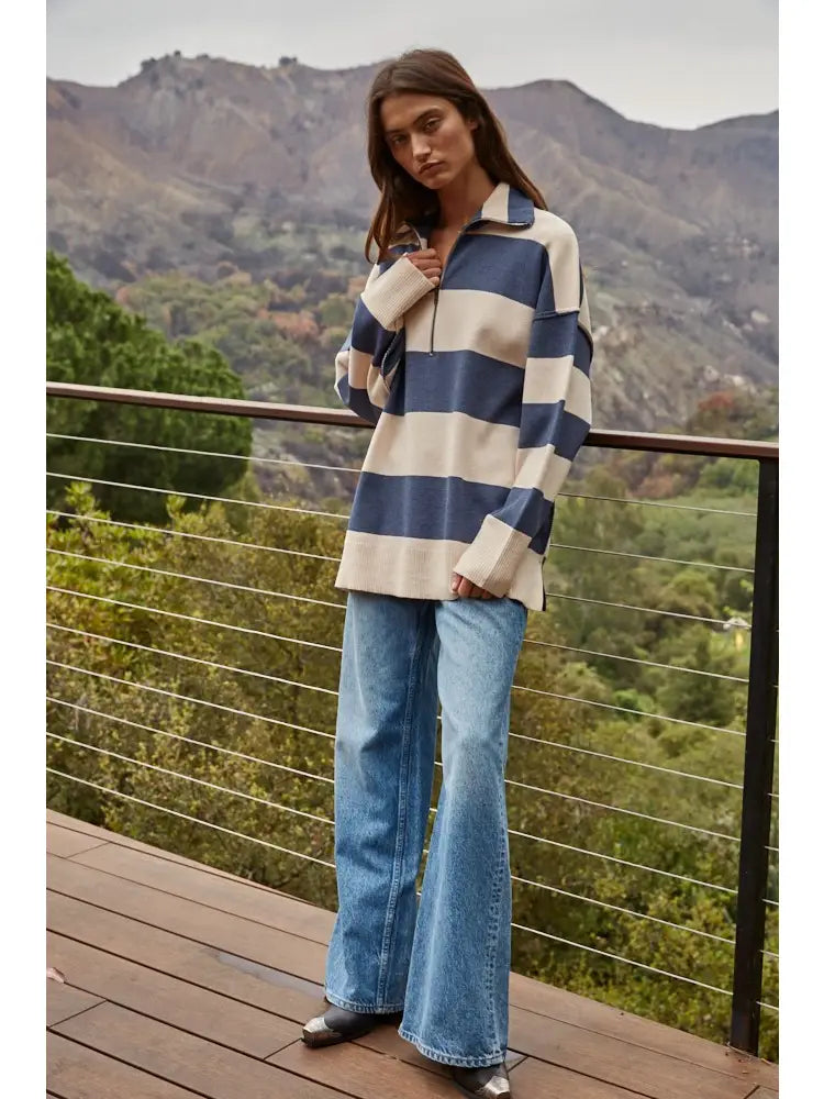 A woman stands on a wooden deck wearing a blue and cream striped long-sleeve quarter-zip pullover and light-wash blue flare jeans, with a mountain landscape in the background.