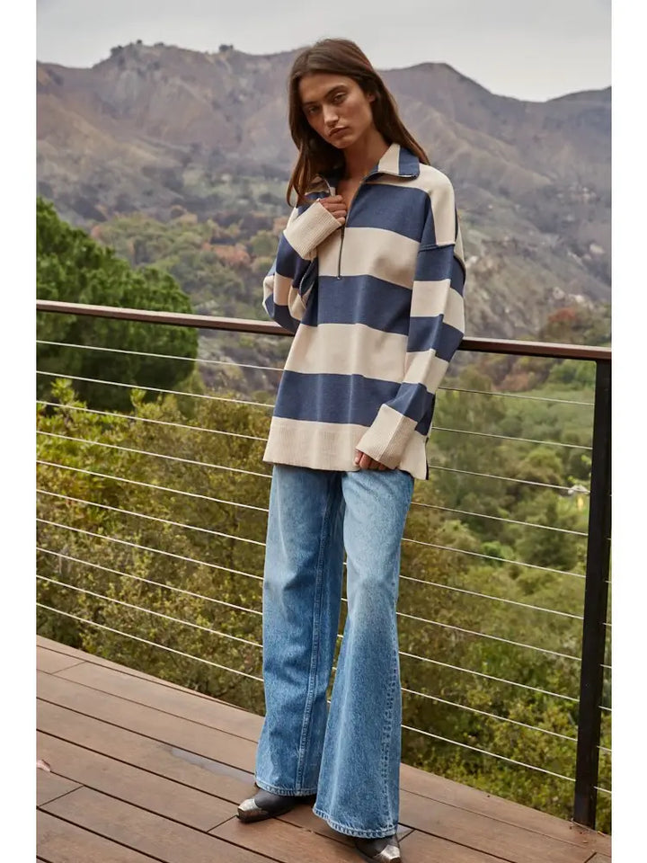 A woman stands on a wooden deck wearing a blue and cream striped long-sleeve quarter-zip pullover and light-wash blue flare jeans, with a mountain landscape in the background.