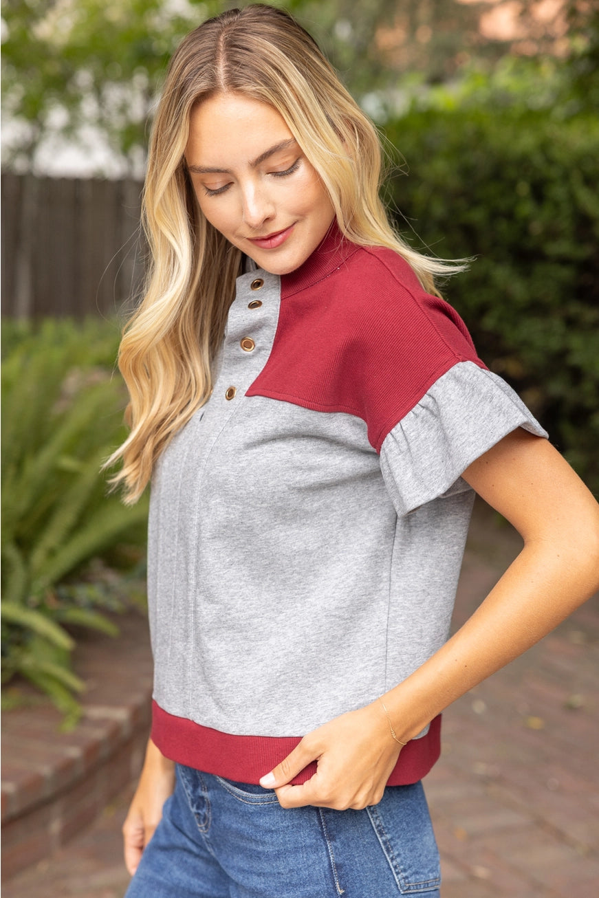 A woman wearing a heather gray short-sleeve top with contrasting burgundy yoke and hem, featuring a buttoned placket, paired with blue jeans in an outdoor garden setting.