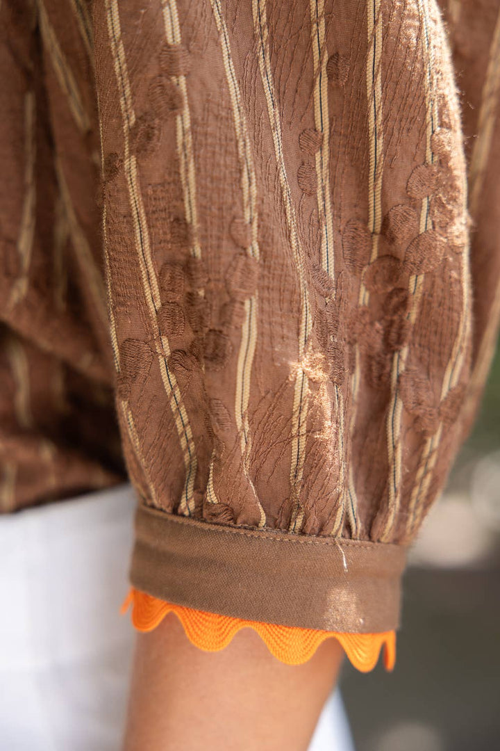 Close-up of a brown sheer embroidered blouse with vertical beige stripes and a bright orange scalloped trim at the gathered elbow cuff.