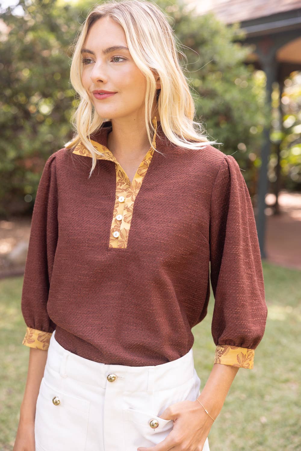 A woman wearing a rust-brown long-sleeve knit top with a gold patterned collar and button placket, paired with white pants, standing outdoors in a garden setting.