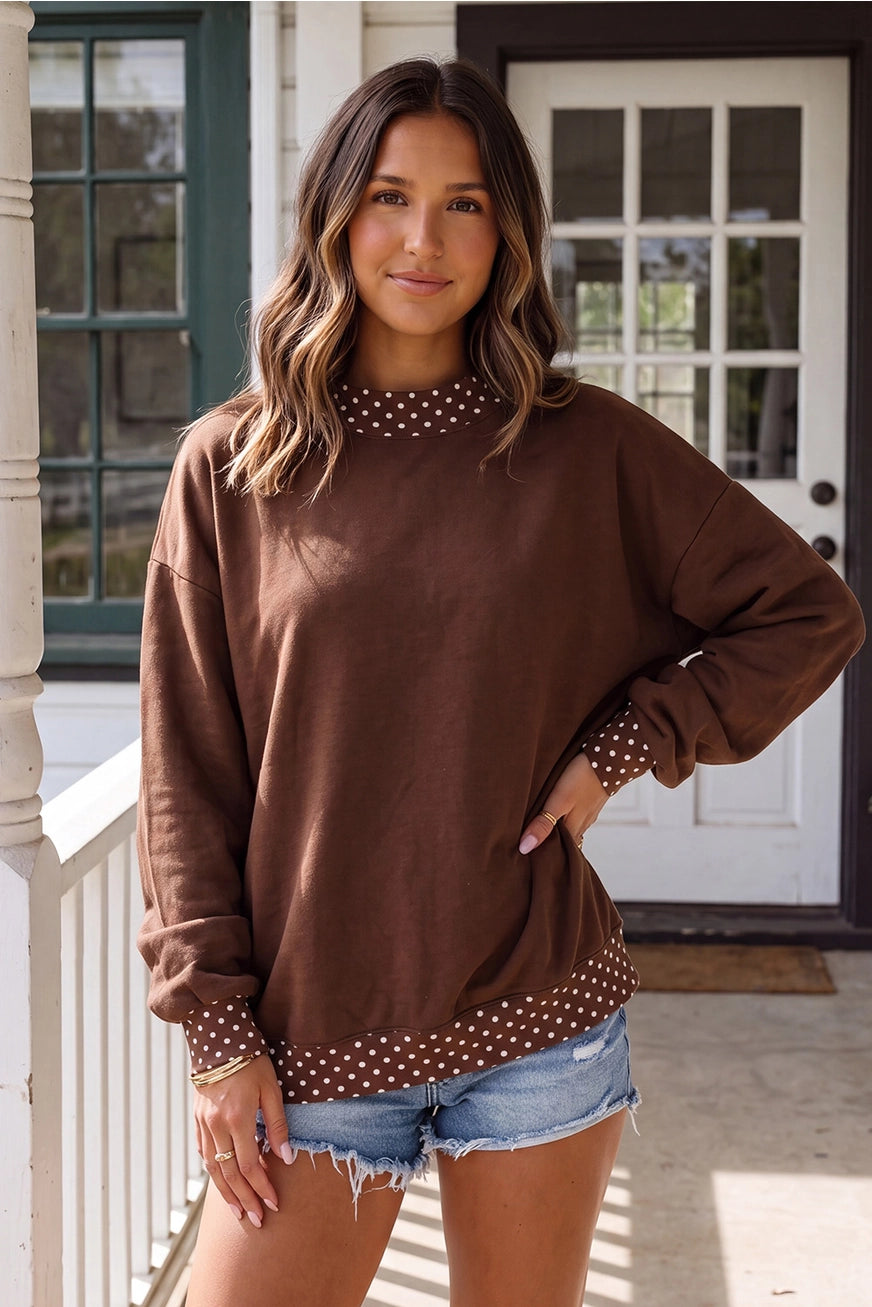A woman modeling a brown long-sleeve sweatshirt with white polka dot trim at the mock neck, cuffs, and hem, paired with denim shorts, standing on a porch.