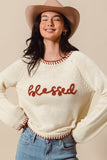 Woman wearing a cream sweater with 'blessed' text, blue jeans, and a beige hat on a beige background