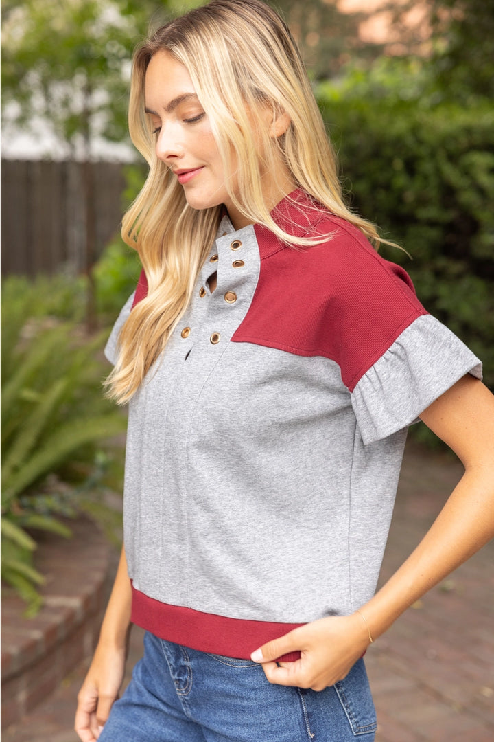 A woman wearing a color-block short-sleeve henley top with a heather gray body and sleeves, and a deep red collar, cuffs, and hem, paired with blue jeans in an outdoor garden setting.