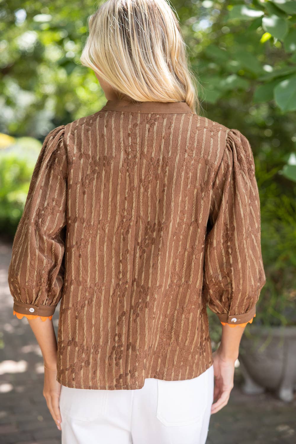 A woman viewed from behind wearing a brown long-sleeve blouse with vertical textured stripes and white pants, set against a blurred green garden background.