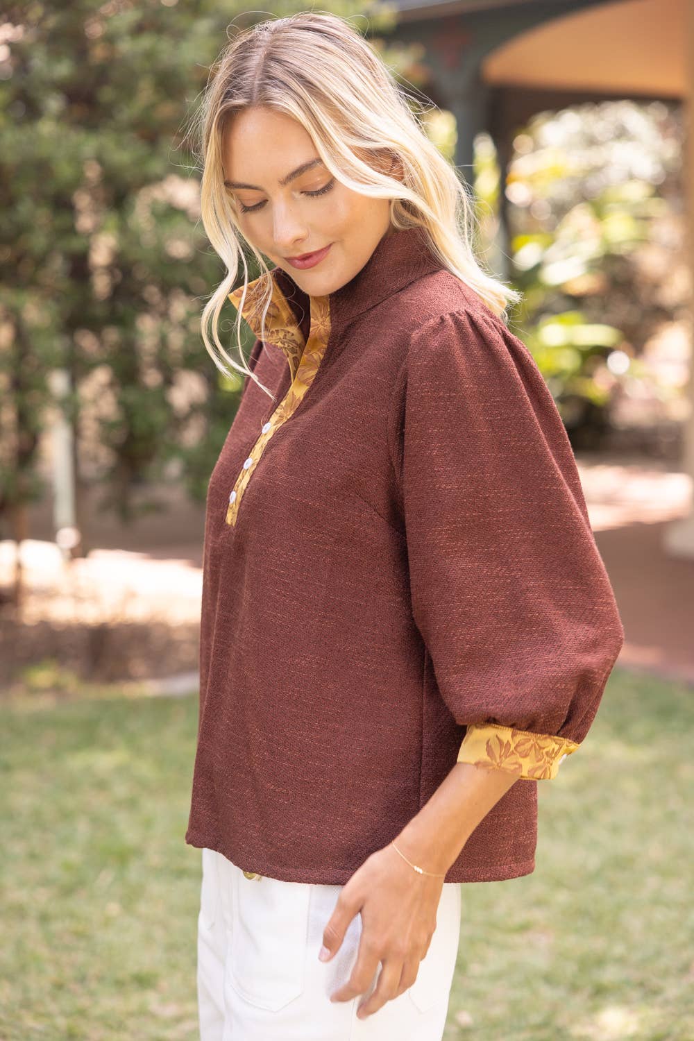 A woman wearing a rust-brown linen blouse with gold lace trim on the collar and cuffs, paired with white pants, standing outdoors in a garden setting.