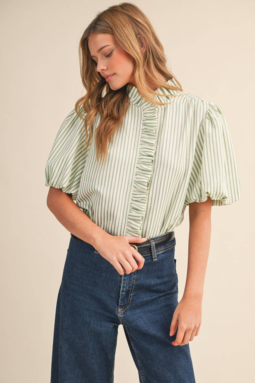 Woman wearing a green and white striped blouse with ruffled sleeves and blue jeans on a beige background