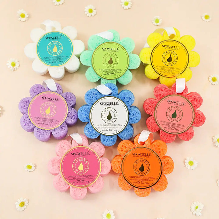 Seven flower-shaped bath sponge bath bombs by Syncellie in assorted pastel and bright colors, arranged in a circular pattern on a light beige background decorated with small white daisy flowers.