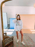 Person taking a mirror selfie wearing a colorful sweatshirt and shorts in a room with a large mirror and colorful backdrops.