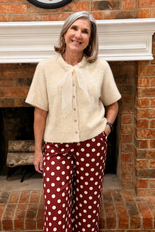 A woman stands in front of a brick fireplace wearing a cream short-sleeve fuzzy knit top with a bow tie neckline and burgundy wide-leg pants with white polka dots.