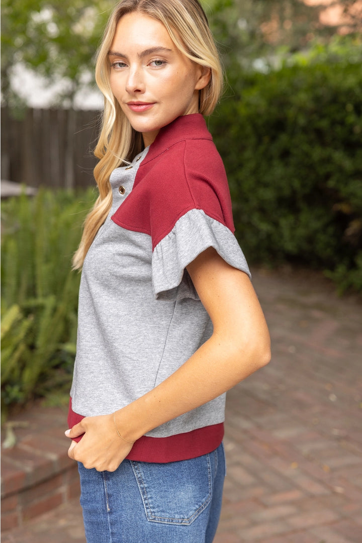 A woman wearing a short-sleeve polo shirt with a heather gray body and contrasting burgundy yoke and hem, paired with blue jeans, standing outdoors on a brick path with green foliage in the background.