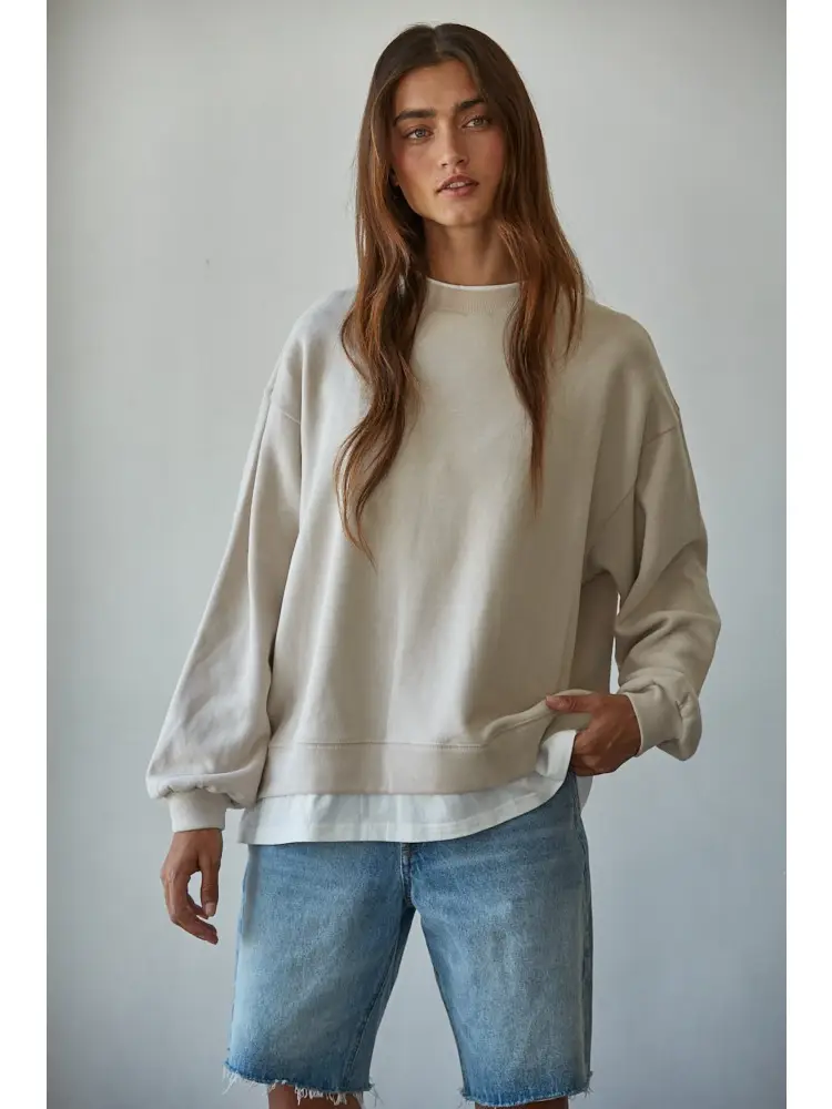 A woman modeling an oversized cream-colored crewneck sweatshirt paired with light-wash denim shorts against a plain light gray background.