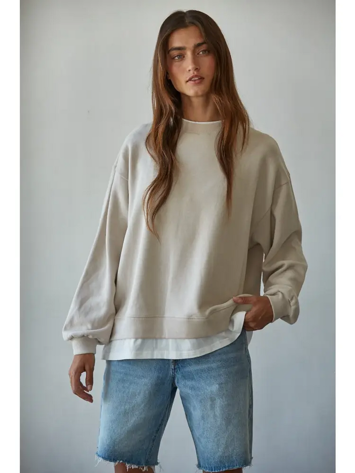 A woman modeling an oversized cream-colored crewneck sweatshirt paired with light-wash denim shorts against a plain light gray background.