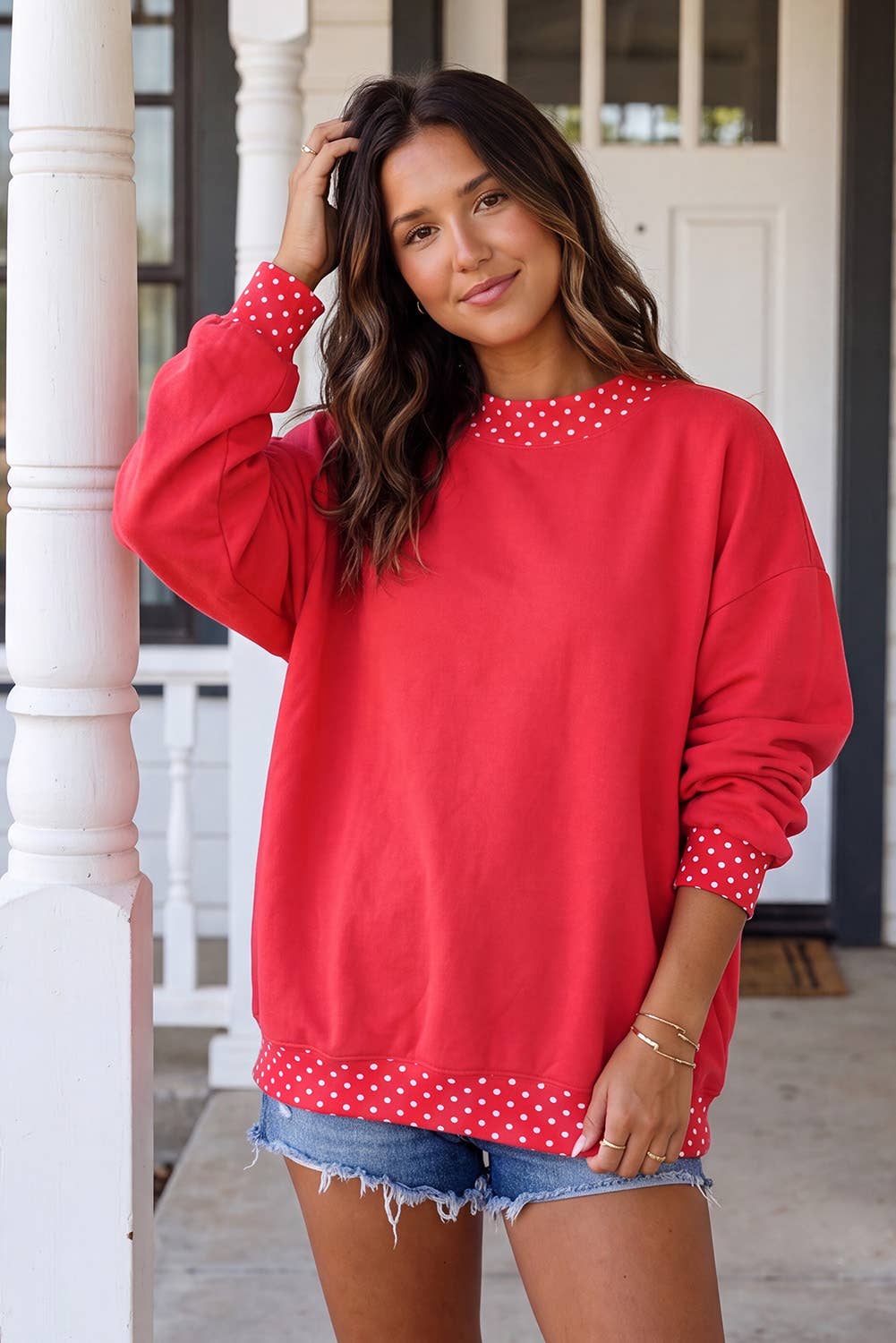 A woman wearing a red crewneck sweatshirt with white polka dot trim on the collar and cuffs, paired with denim shorts, standing on a porch.