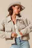 Woman wearing a beige quilted jacket, white top, and light blue jeans with a neutral background