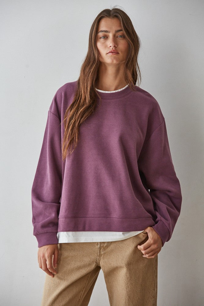 A woman wearing a dusty purple crewneck sweatshirt over a white t-shirt and beige pants, standing against a neutral gray background.