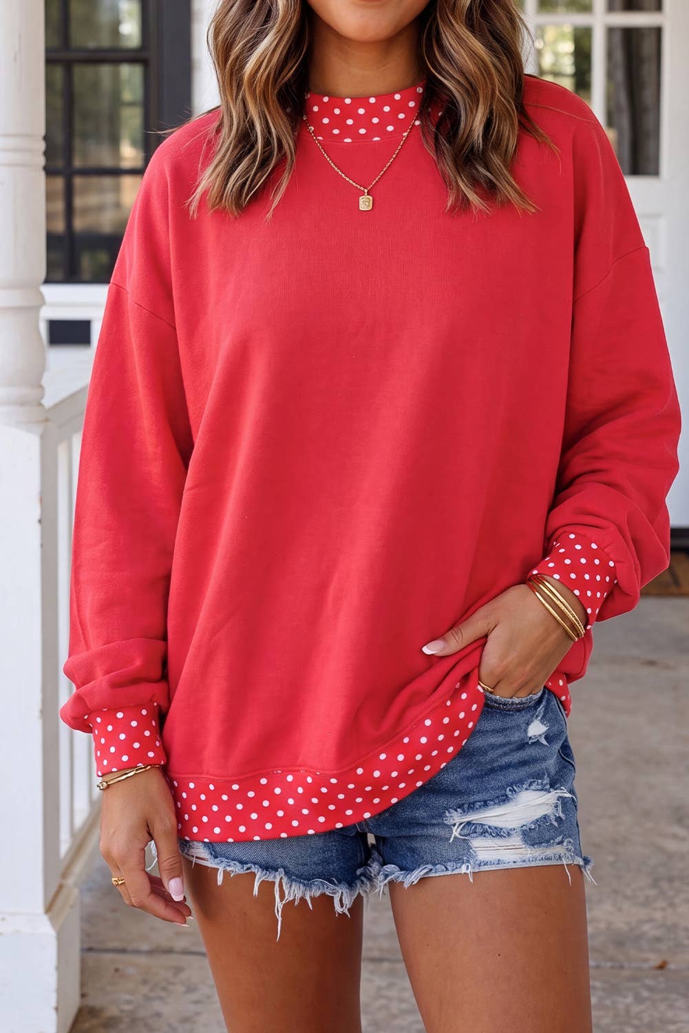 A woman wearing a red long-sleeve sweatshirt with white polka dot trim on the collar, cuffs, and hem, paired with distressed denim shorts.