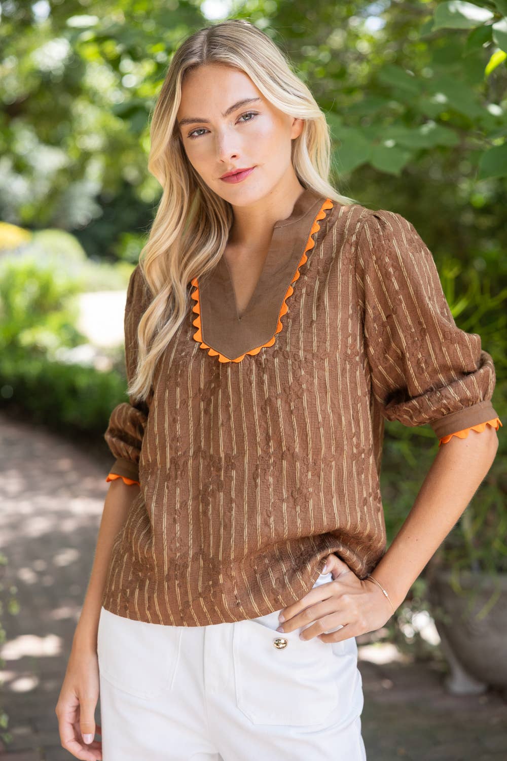 A woman wearing a brown and beige vertically striped blouse with orange scalloped trim on the V-neck and cuffs, paired with white pants, standing outdoors in front of a blurred green garden background.