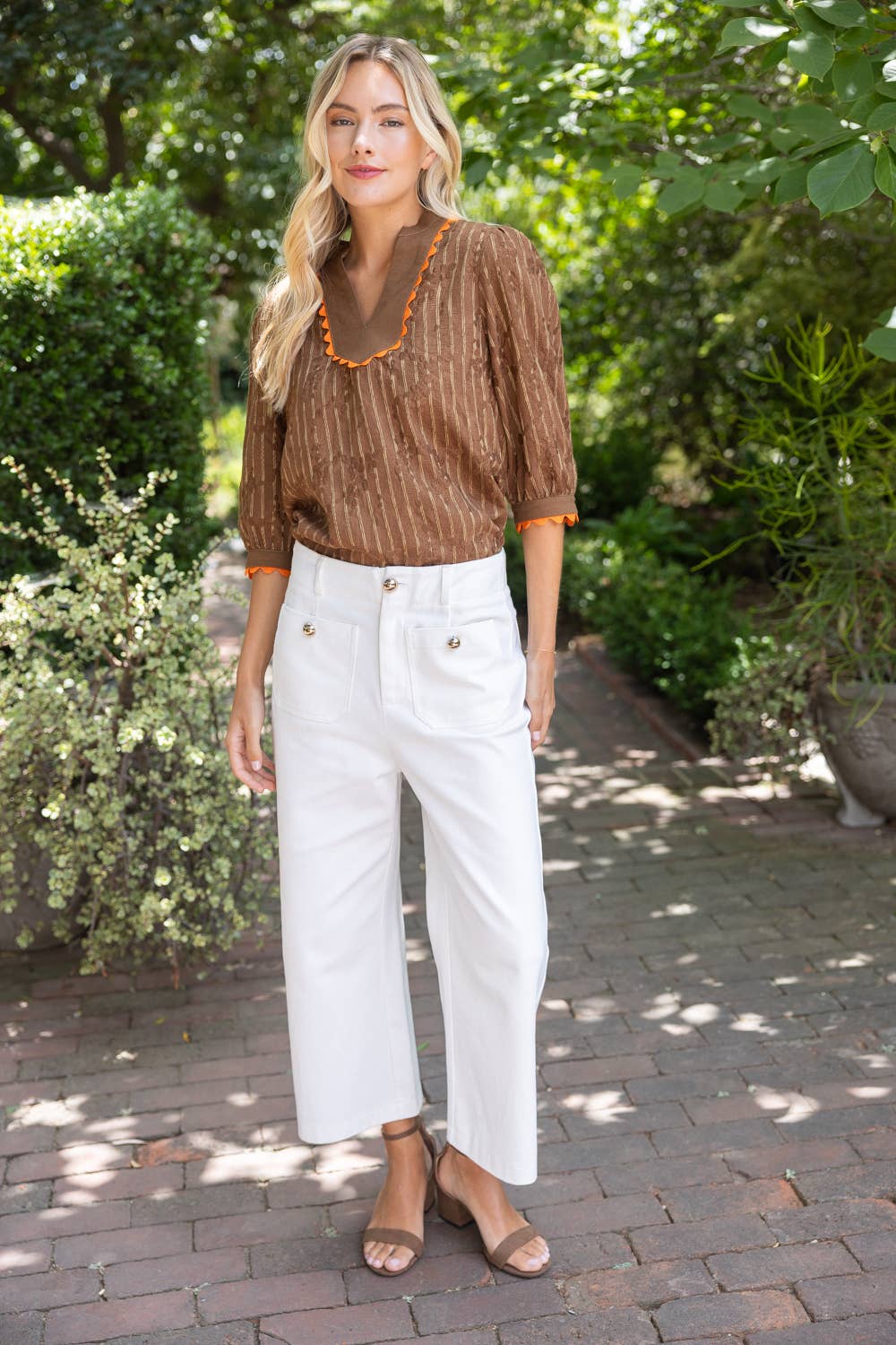 A woman standing outdoors on a brick path surrounded by green foliage, wearing a brown textured blouse with orange trim, white cropped pants, and tan heeled sandals.