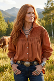 Woman in a rust-colored blouse and blue jeans standing in a field with mountains in the background