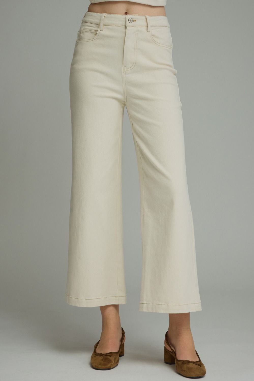 A person wearing off-white cropped wide-leg jeans with brown slingback flats, shown from the waist down against a light gray studio background.