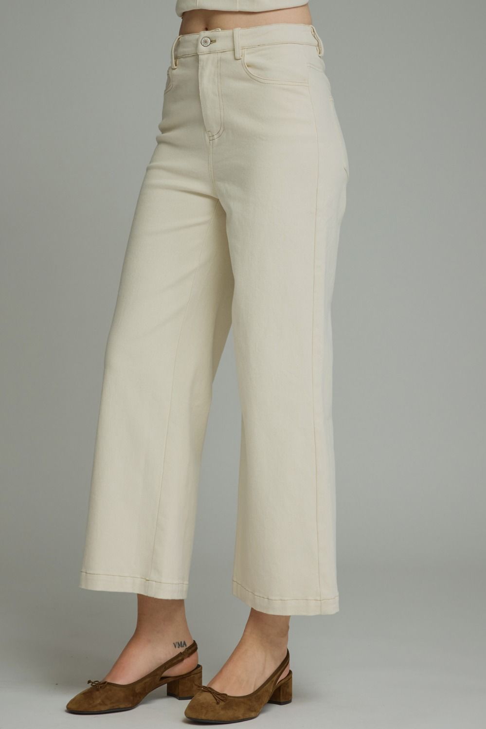 A model is wearing a pair of off-white cropped wide-leg jeans with brown slingback heels, shown from the waist down against a neutral gray studio background.