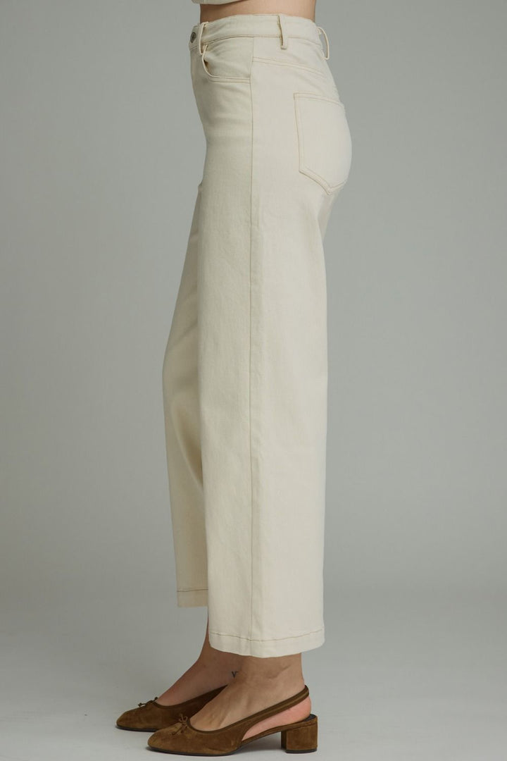 A side profile view of a person wearing off-white cropped wide-leg jeans and brown slingback block-heel mules against a neutral gray studio background.