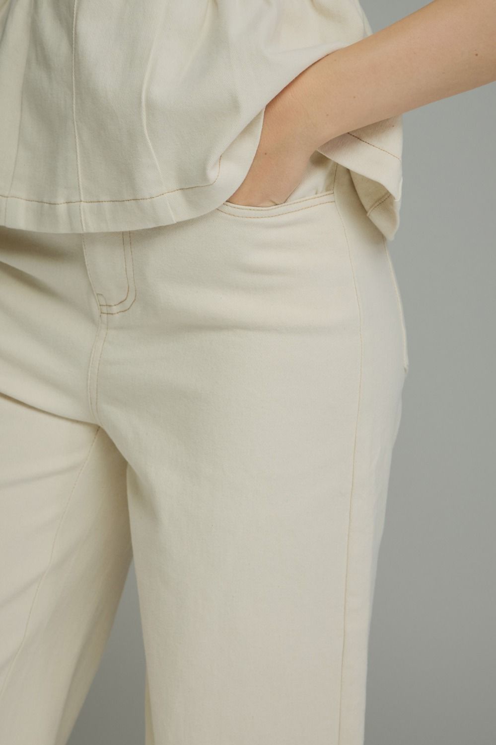 Close-up of a person wearing cream-colored trousers and a matching top, with one hand in a pocket, against a plain light gray background.