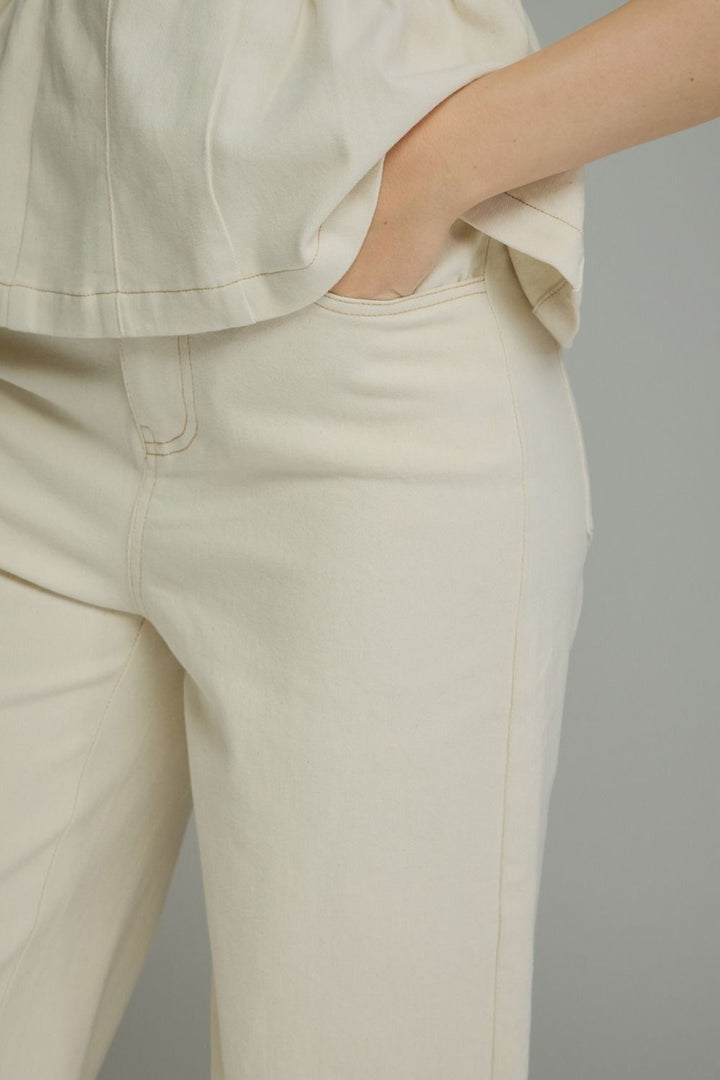 Close-up of a person wearing cream-colored trousers and a matching top, with one hand in a pocket, against a plain light gray background.