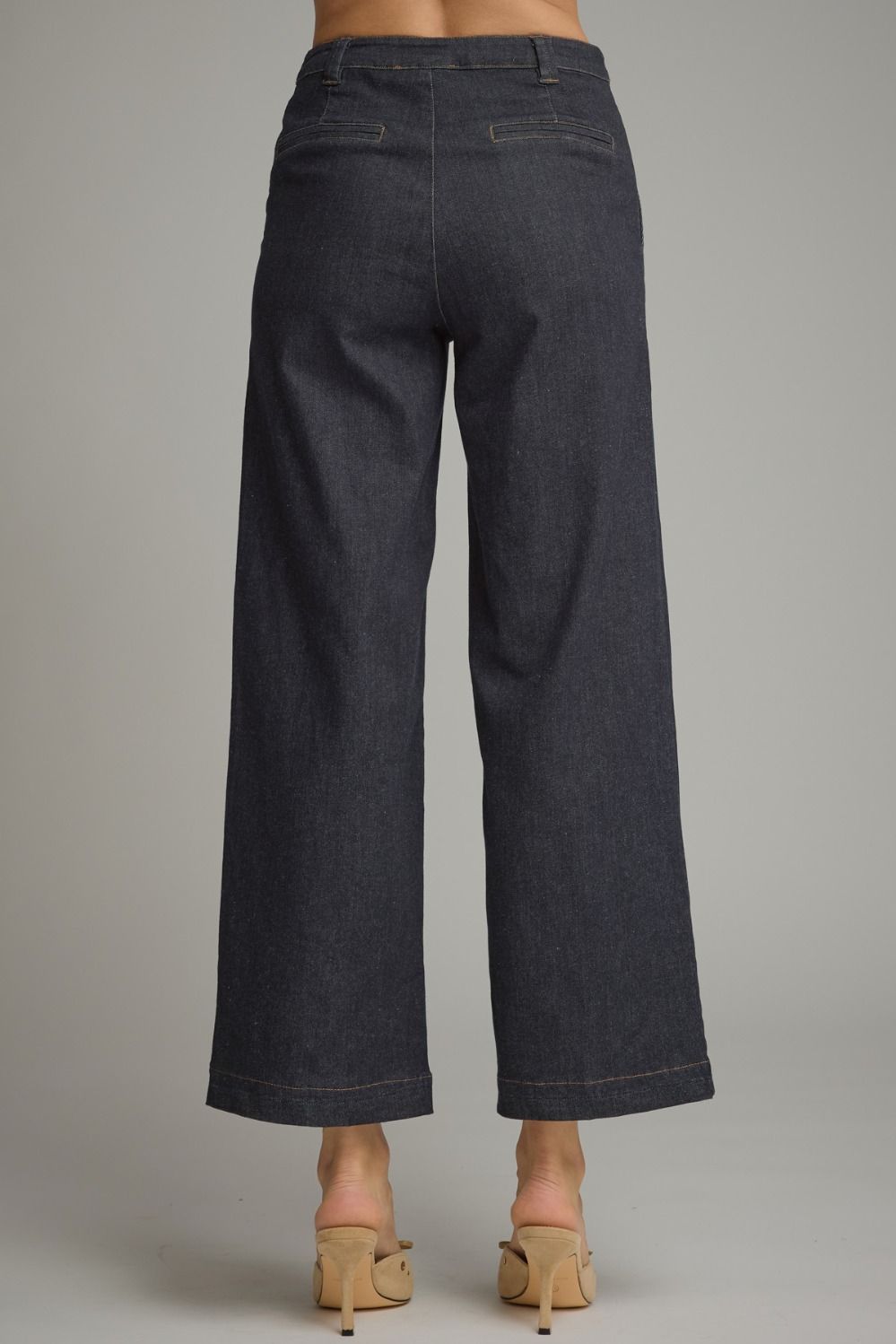 Rear view of a person wearing dark indigo wide-leg cropped jeans with beige mule heels, shown against a plain light gray studio background.