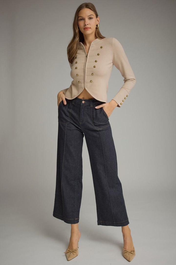 A model stands against a light gray studio background wearing a beige cropped long-sleeve jacket with gold buttons, dark wide-leg cropped jeans, and beige pointed flats.