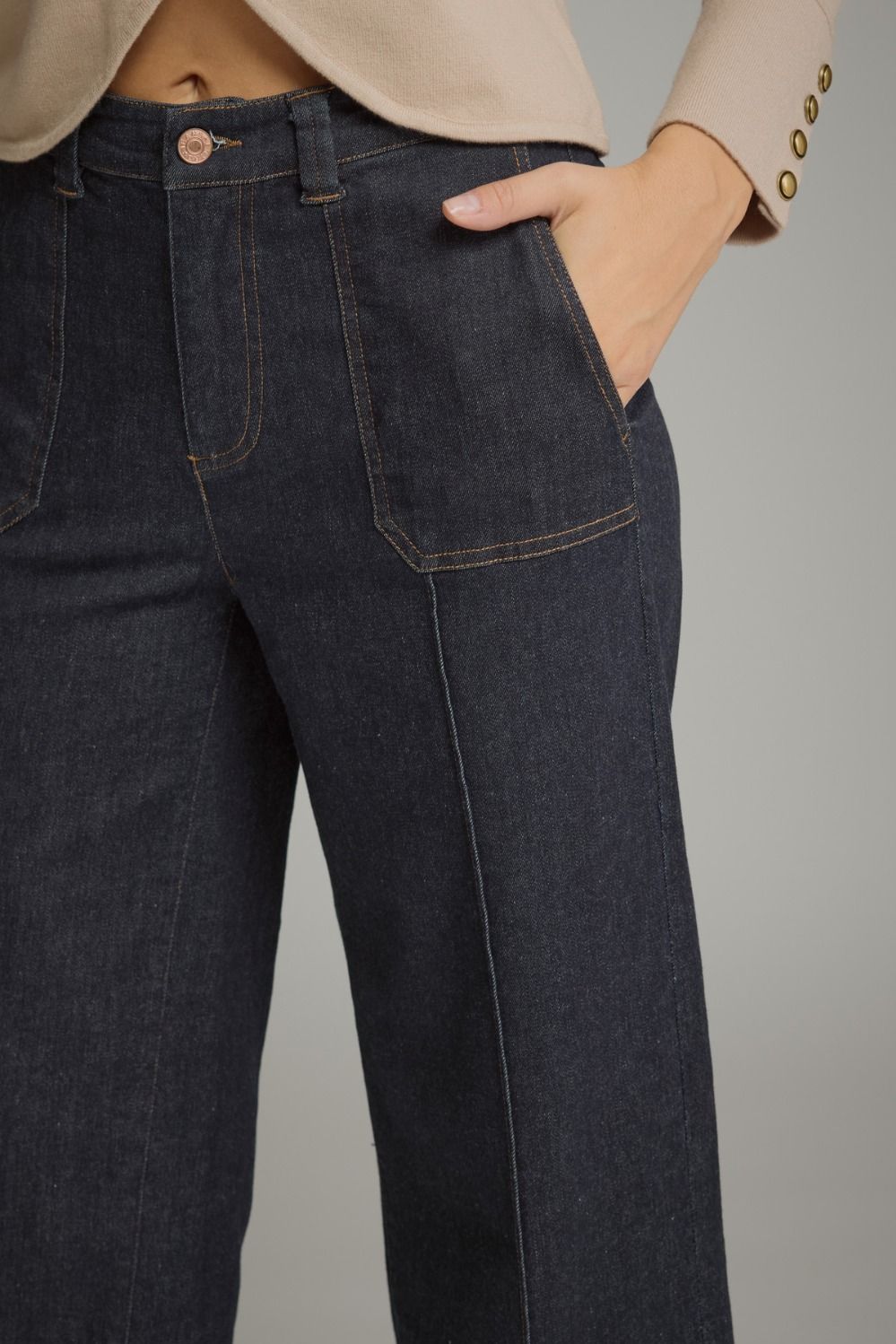 Close-up of a person wearing dark indigo denim jeans with a beige blazer, showing the side pocket, front seam, waistband, and hand in the pocket against a plain gray background.
