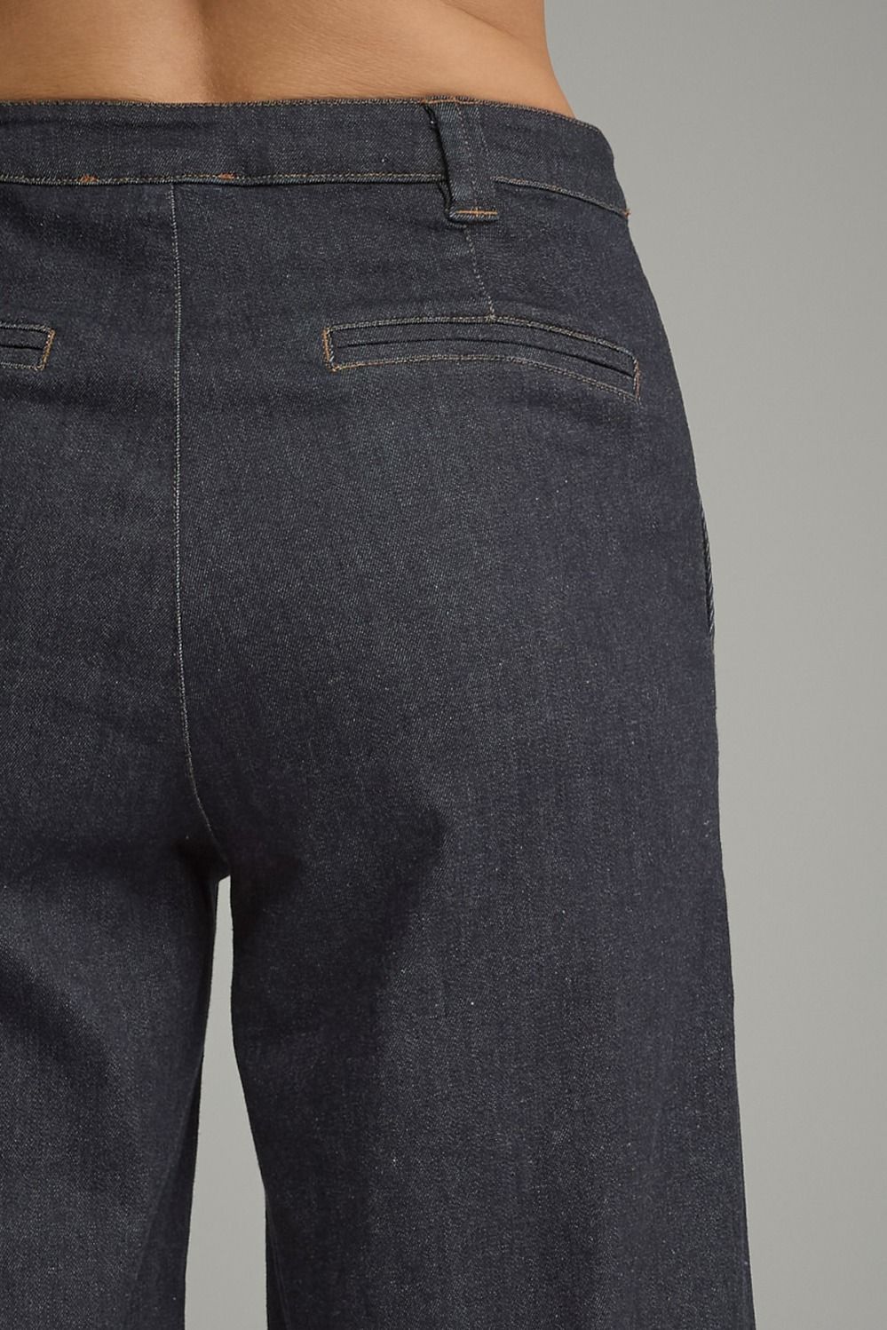 Close-up back view of a person wearing dark indigo denim jeans, showing the waistband, belt loops, back pockets, and fabric texture against a plain gray background.