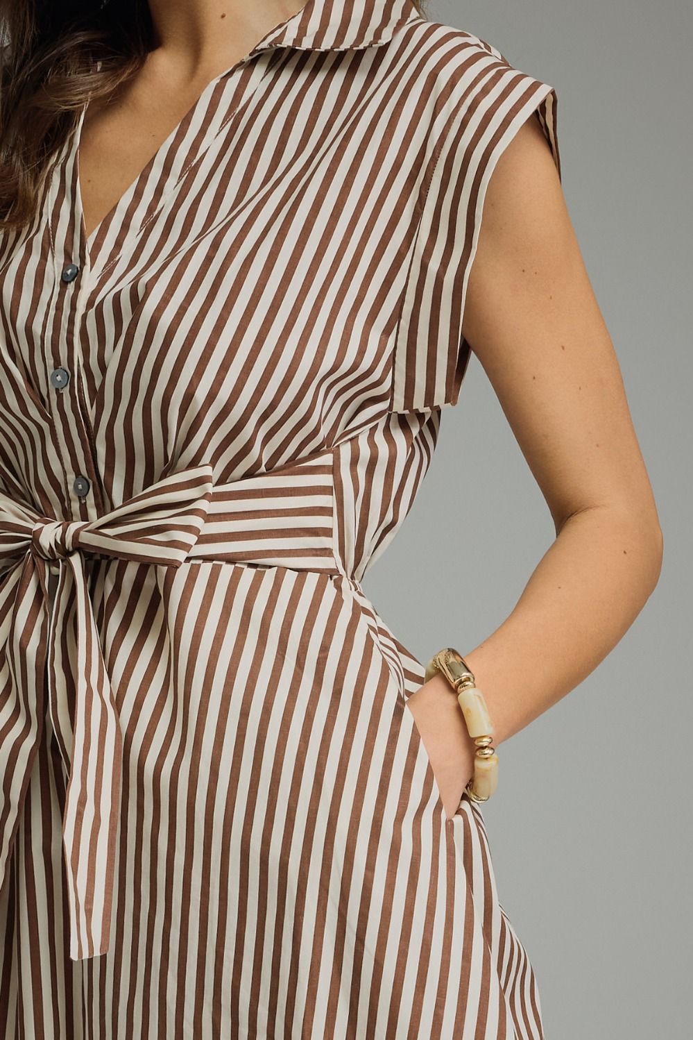 Close-up of a woman wearing a sleeveless collared shirt dress with vertical brown and off-white stripes, a button front, and a self-tie waist belt. She is wearing a gold-toned bracelet and has one hand in her pocket against a solid gray background.
