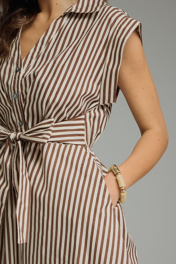 Close-up of a woman wearing a sleeveless collared shirt dress with vertical brown and off-white stripes, a button front, and a self-tie waist belt. She is wearing a gold-toned bracelet and has one hand in her pocket against a solid gray background.