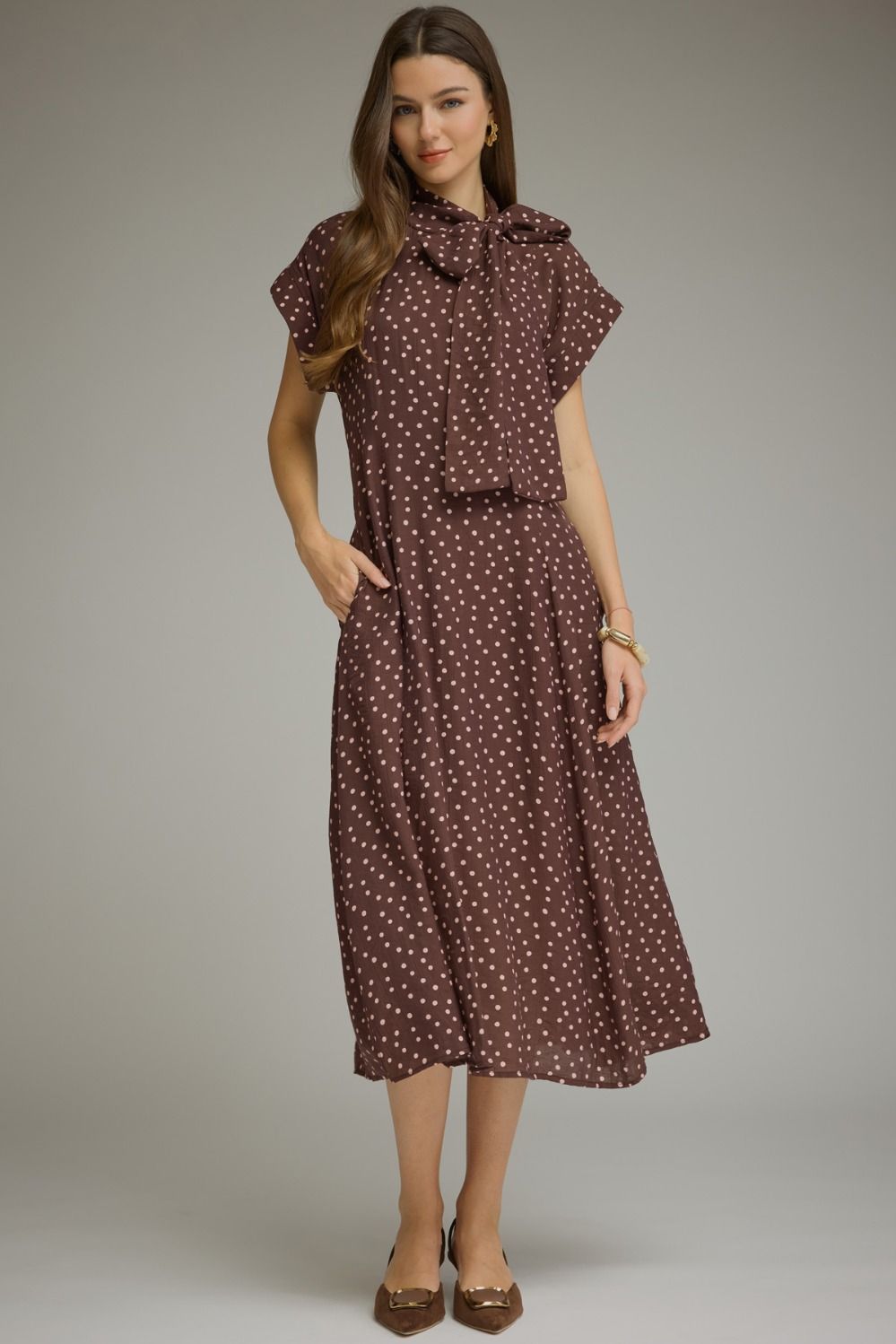 A woman modeling a brown midi dress with a white polka dot pattern, featuring short flutter sleeves, a ruffled collar, and a matching fabric tie belt at the waist, set against a plain gray studio background.
