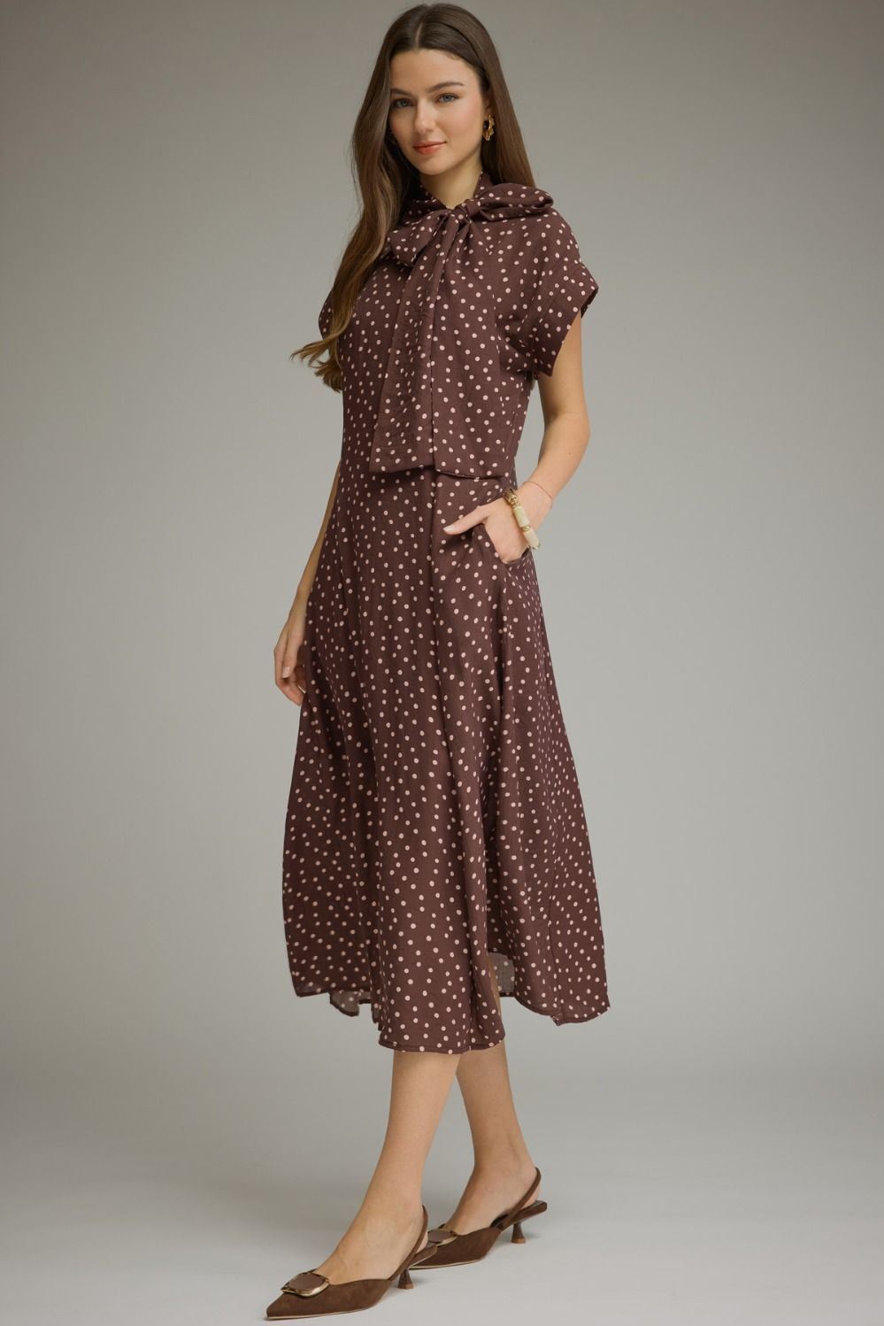 A woman modeling a brown midi dress with a white polka dot pattern, featuring short flutter sleeves, a ruffled tie-neck collar, and a matching self-tie belt, set against a plain gray studio background.