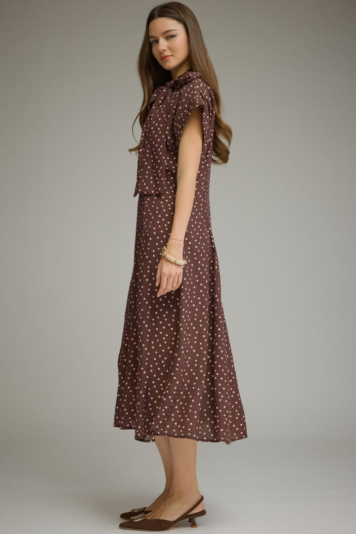 A woman modeling a brown midi dress with a white polka dot pattern, featuring a high neck, ruffled cap sleeves, and a matching belt, set against a plain gray studio background.