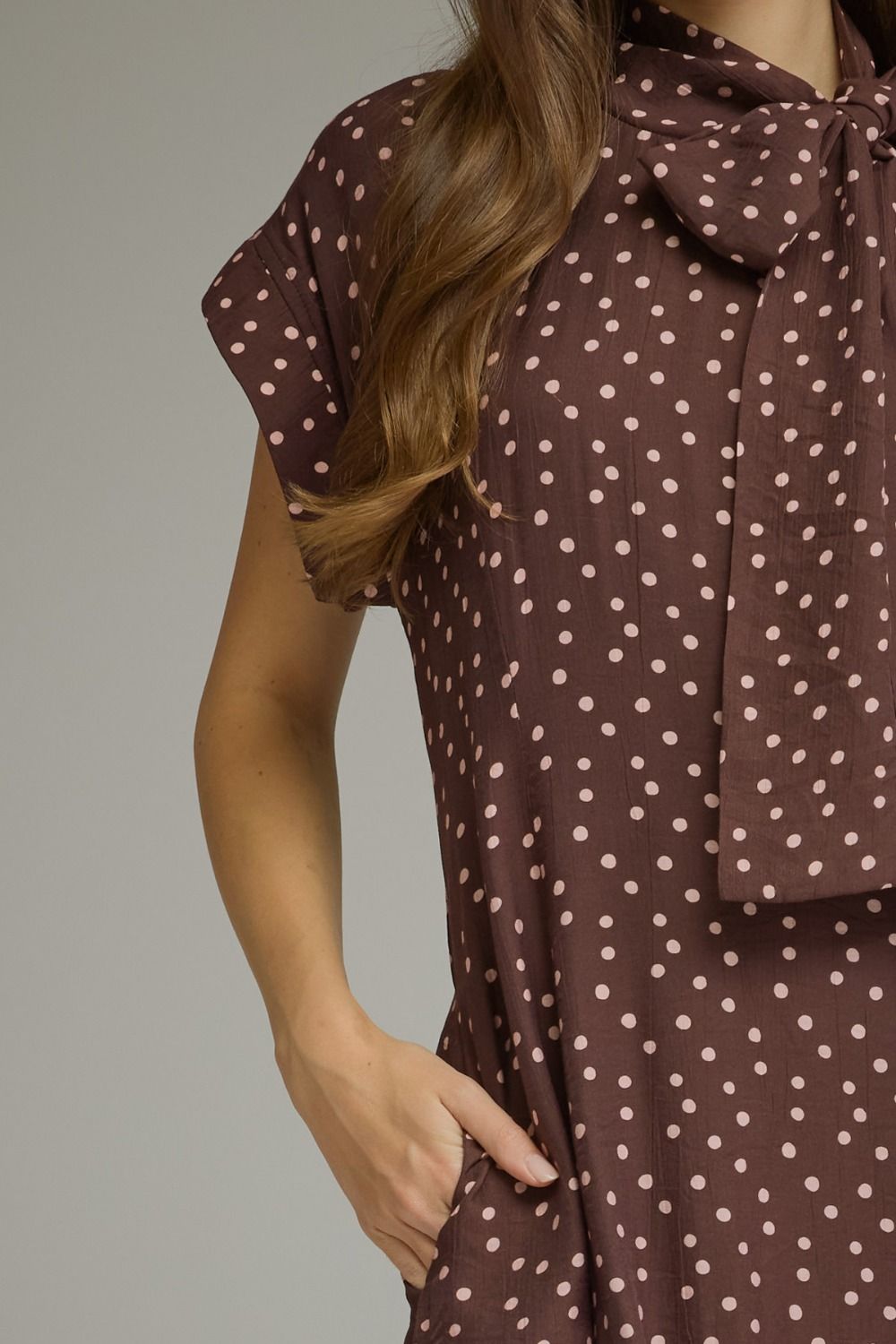 A close-up of a woman wearing a brown short-sleeve dress with a white polka dot pattern and a large bow tie at the collar, set against a plain gray background.