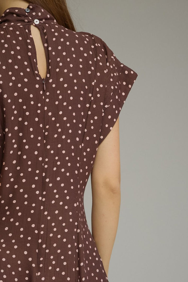 Close-up of a person wearing a brown dress with a white polka dot pattern, showing the back of the dress with a keyhole cutout and short flutter sleeves against a plain gray background.