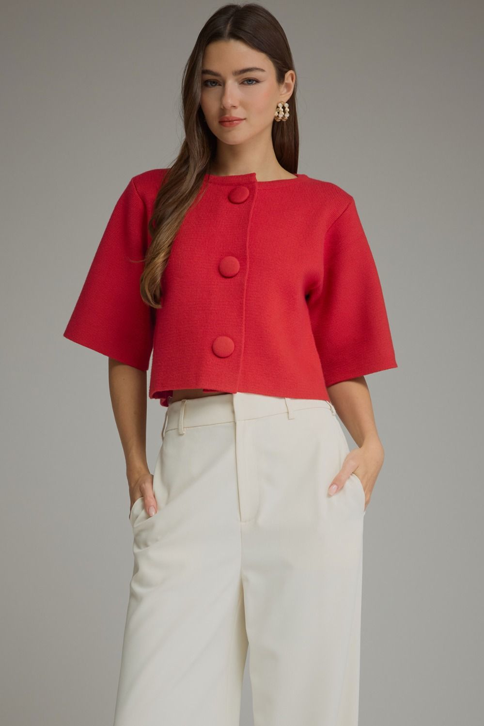 A woman modeling a bright red cropped short-sleeve jacket with three large front buttons, paired with white wide-leg trousers, against a solid gray studio background.