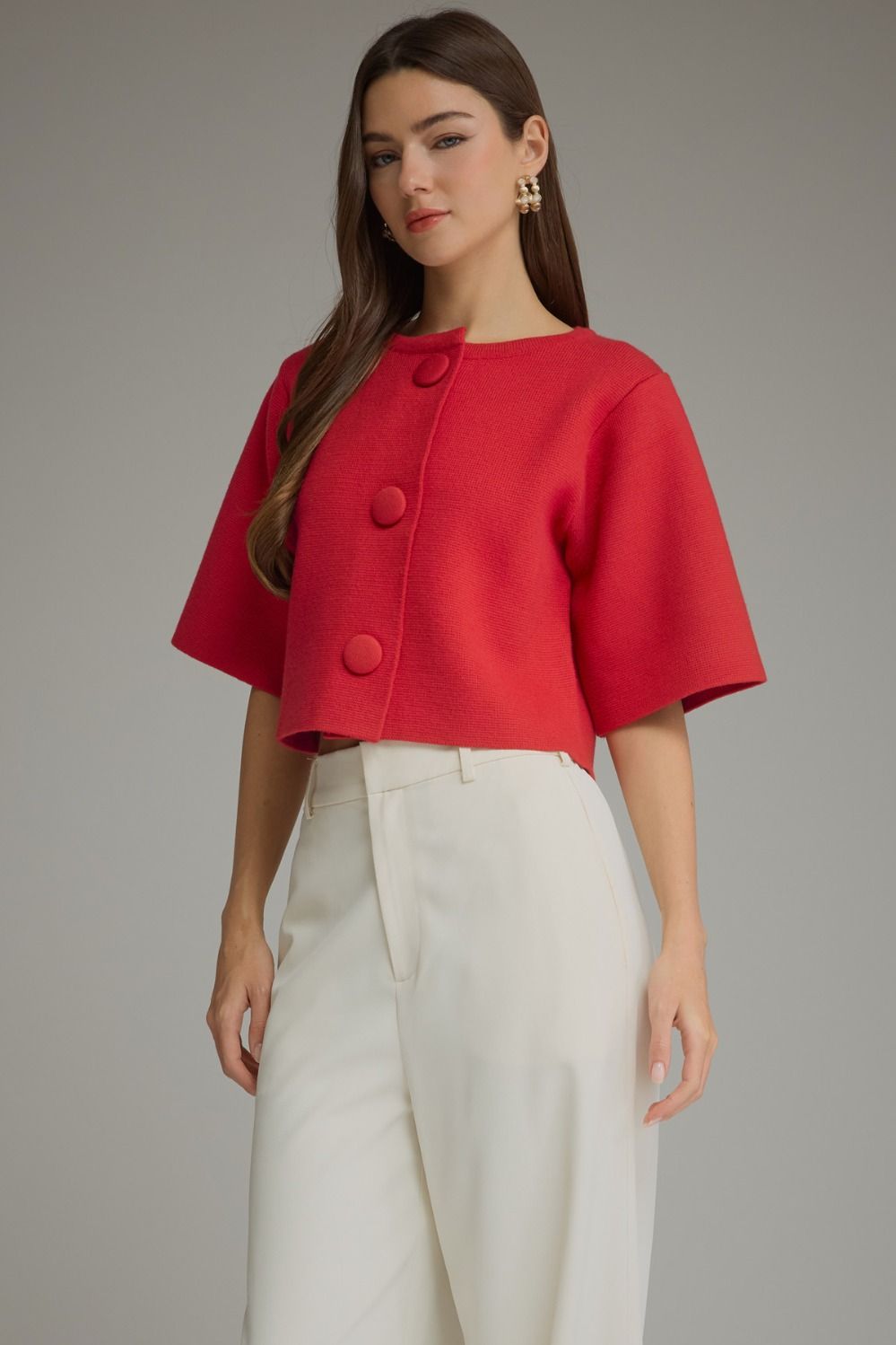 A woman modeling a bright red cropped short-sleeve jacket with large decorative buttons, paired with white trousers, against a neutral gray studio background.
