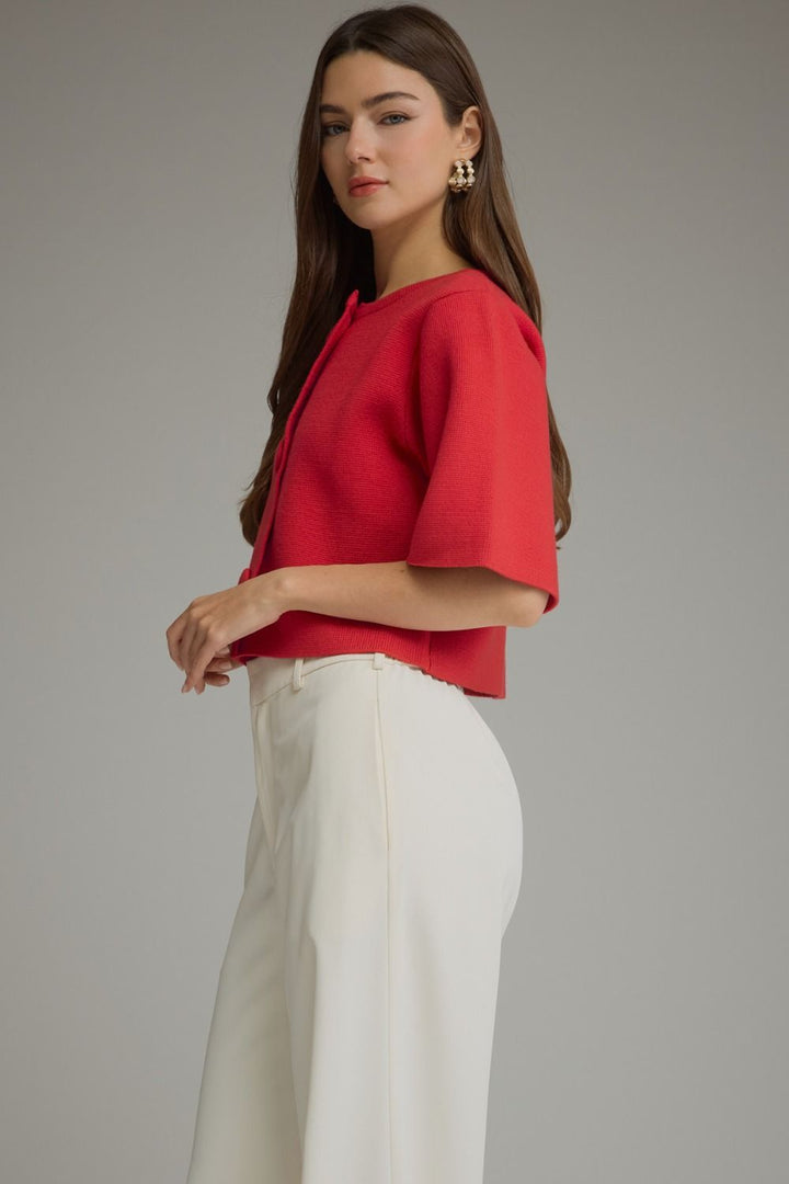 A woman modeling a bright red short-sleeve cropped top with a round neckline, paired with white wide-leg trousers, against a solid gray studio background.