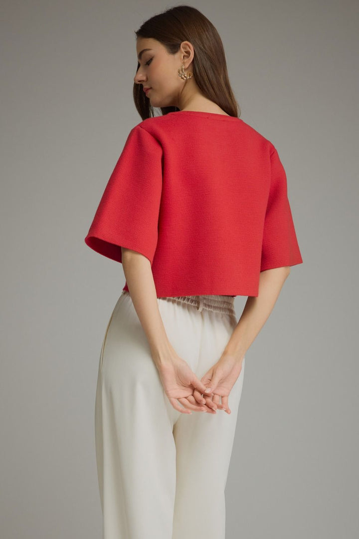 A woman shown from the back wearing a bright red short-sleeve cropped top and off-white trousers, standing against a plain light gray studio background.