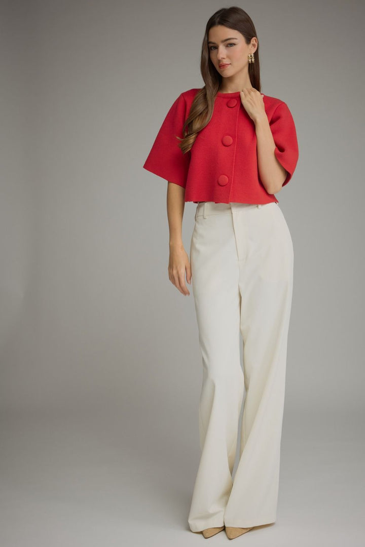 A woman modeling a bright red short-sleeve cropped jacket with large decorative buttons, paired with high-waisted off-white wide-leg trousers, against a neutral gray studio background.