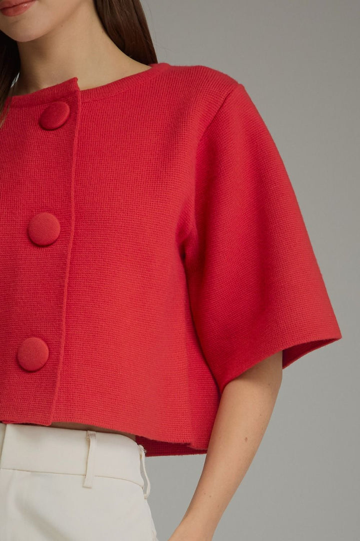 A close-up of a model wearing a vibrant red cropped knit top with three large matching buttons down the front, paired with white trousers against a neutral gray background.