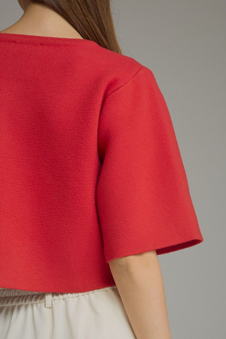 Close-up back view of a person wearing a bright red short-sleeve knit top with a wide neckline and textured fabric, paired with light beige bottoms, against a plain gray background.
